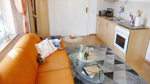 Ein kompakter Wohnbereich mit Sofa, Glastisch, rundem Tisch, Küchenzeile mit Herd und Spüle sowie einer Garderobe.
