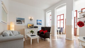 Ein modernes Wohnzimmer mit einem grauen Sofa, einem schwarzen Sessel und einem weißen Couchtisch. Ein Fernseher und eine kleine Kommode mit einem Blumenstrauß sind ebenfalls vorhanden.