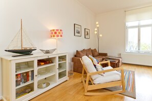A living room with a wooden rocking chair, a white cabinet with glass doors, a small sailboat model, and a lamp with a floral lampshade. - Villa Charlottes Höh, app. 3 (Heringsdorf)