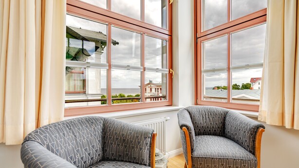 Een kamer met twee gestoffeerde fauteuils, een radiator en grote ramen met uitzicht op gebouwen en groen.
