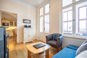 Ein modernes Wohnzimmer mit einer Couch, einem Couchtisch und einem Sessel. Im Hintergrund ist eine Küchenzeile mit Schränken und einer Mikrowelle zu sehen.