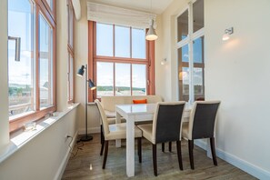 Un espace repas avec une table et des chaises blanches, de grandes fenêtres et une vue sur la ville.