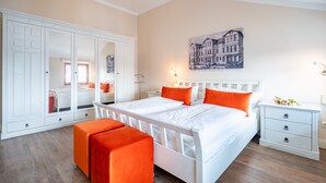 Une chambre avec un lit blanc, des coussins orange, une table de chevet, un miroir et un tableau accroché au mur.
