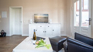 Une pièce avec un meuble blanc, un téléviseur accroché au mur, une bouteille de vin, des verres et un bouquet de fleurs sur une table.