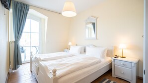 Een tweepersoonsbed met wit beddengoed, een nachtkastje met een lamp, een spiegel aan de muur en een raam met gordijnen.