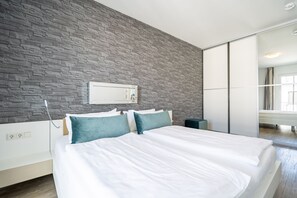 A modern bedroom with a stone-textured wall, a large bed with white bedding and teal pillows, a bedside table, and a mirror. - Dünenpalais, App. 19 (Heringsdorf)
