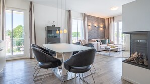 Un salon moderne avec une cheminée, un coin repas doté d’une table en verre et une cuisine agrémentée d’un mur en pierre.