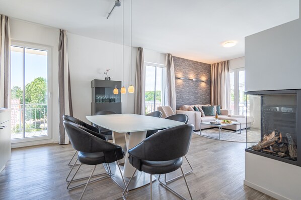 A modern living room with a fireplace, a dining area with a glass table, and a kitchen with a stone accent wall. - Dünenpalais, App. 19 (Heringsdorf)