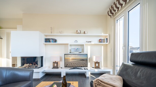 Ett modernt vardagsrum med en öppen spis, en platt-TV, ett soffbord med en vinflaska och blommor samt en svart lädersoffa.
