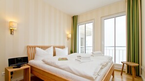 A neatly made bed with white linens, a bedside table with a flat-screen TV, and a window with green curtains.