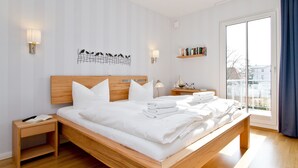 A modern bedroom with a wooden bed, white bedding, a nightstand, wall-mounted lamps, a decorative wall piece, a bookshelf, and a large window with a view.