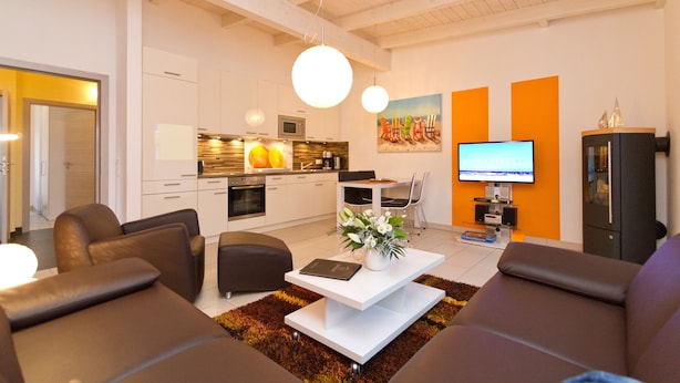 Una sala de estar moderna con sofá, mesa de centro y televisor. Hay una zona de cocina con armarios y microondas.