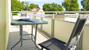 Un balcón con una mesa y silla de metal, con vista a una zona residencial con árboles y casas.