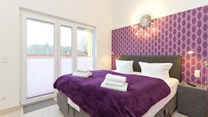 Un dormitorio moderno con una cama grande, una colcha morada y almohadas blancas. Hay una mesita de noche con un control remoto y una lámpara de pared.