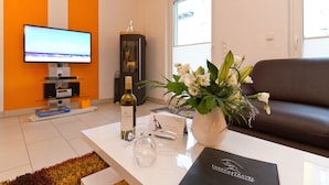 Una sala moderna con un televisor de pantalla plana, una botella de vino, copas y un florero sobre una mesa de centro blanca.