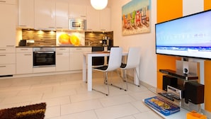 Una cocina moderna con armarios blancos, un microondas y una cafetera. Hay un comedor con una mesa y sillas. Un televisor está montado en la pared, y se aprecia un cuadro.