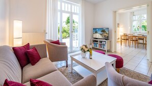 Ett modernt vardagsrum med en soffa, ett soffbord och en tv som står på ett tv-bord. I bakgrunden syns en matplats med ett bord och stolar genom en öppen dörr.