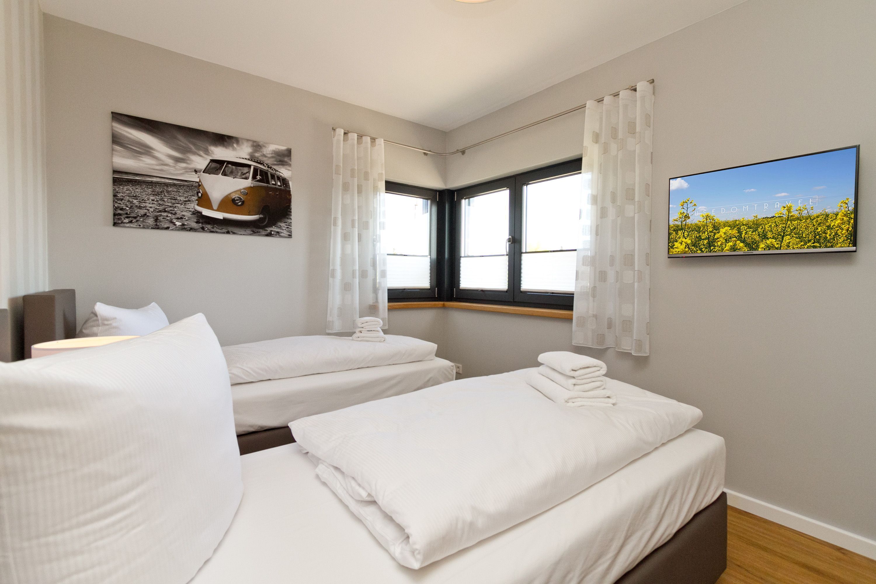 Een hotelkamer met twee bedden, een groot raam met gordijnen, een schilderij van een busje, en een televisie aan de muur.