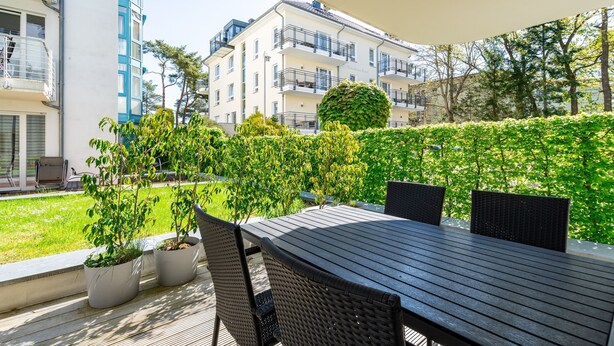 An outdoor patio area with a wooden table and chairs, surrounded by greenery and a hedge. - Seeresidenz Haus Atlantic, App. C02 (Heringsdorf)