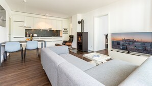 Un saló modern amb un sofà gris, una taula de centre i una televisió que mostra una escena de platja. Es veu un cuina amb armaris blancs i una zona de menjador amb cadires al fons.