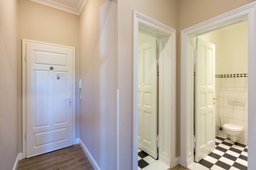 Un couloir avec une porte blanche, un sol à carreaux et une salle de bain visible à travers une porte entrouverte.