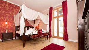 A four-poster bed with white drapes, a wooden nightstand, a red rug, and a large window with red curtains. - Villa Julius, App. 4 Marrakech (Heringsdorf)