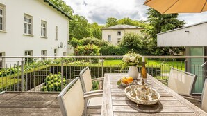 A wooden deck with a table set for dining, surrounded by a metal railing, overlooking a garden and buildings. - Villa Julius, App. 4 Marrakech (Heringsdorf)