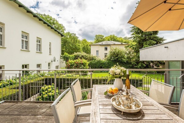 A wooden deck with a table set for dining, surrounded by a metal railing, overlooking a garden and buildings.