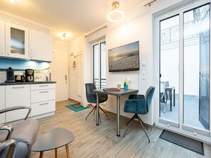 Eine moderne Küche mit weißen Schränken, einer Kaffeemaschine und einem Spülbecken. Es gibt einen Essbereich mit Tisch und Stühlen sowie ein großes Fenster mit Blick auf den Strand.