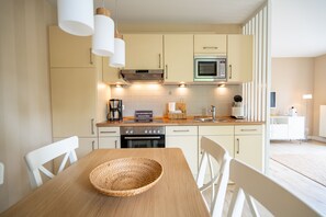 Eine moderne Küche mit hellen Schränken, einer Mikrowelle und einer Kaffeemaschine. Ein Holztisch mit gewebter Tischmatte und weiße Stühle sind ebenfalls vorhanden.