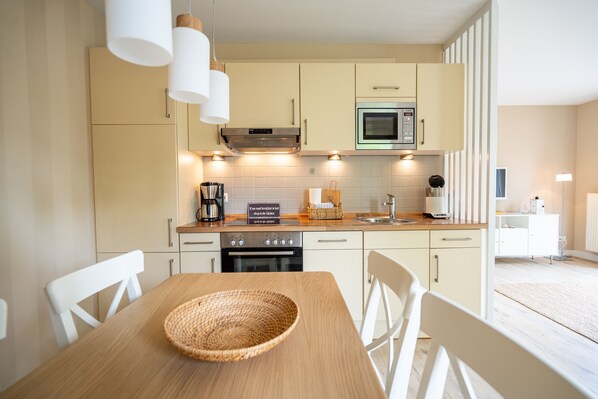 A modern kitchen with light-colored cabinets, a microwave, and a coffee maker. There is a wooden table with a woven placemat and white chairs.