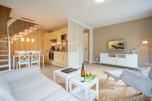 A modern living room with a staircase, white furniture, a dining area, and a kitchen in the background.