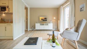 Ein modernes Wohnzimmer mit einem weißen Sofa, einem Esstisch mit Buch und Wein, sowie einer Küchenzeile mit Mikrowelle und Schränken.