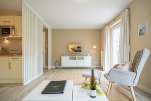 Ein modernes Wohnzimmer mit einem weißen Sofa, einem Esstisch mit Buch und Wein, sowie einer Küchenzeile mit Mikrowelle und Schränken.