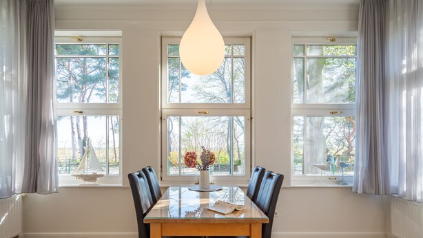 A dining area with a glass table, four chairs, a vase with flowers, and three windows with curtains. - Villa Medici Heringsdorf, App. 07 (Heringsdorf)
