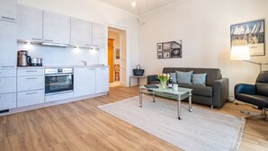 A modern kitchen with white cabinets, a built-in oven, and a coffee maker. There is a glass coffee table with a vase of flowers in the living area, which includes a grey sofa and a black leather chair. - Villa Medici Heringsdorf, App. 07 (Heringsdorf)