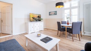 Ein modernes Wohnzimmer mit einem weißen Sofa, einem Couchtisch mit einem Buch, einem Essbereich mit Tisch und Stühlen und Blick auf einen Steg durch ein Fenster.