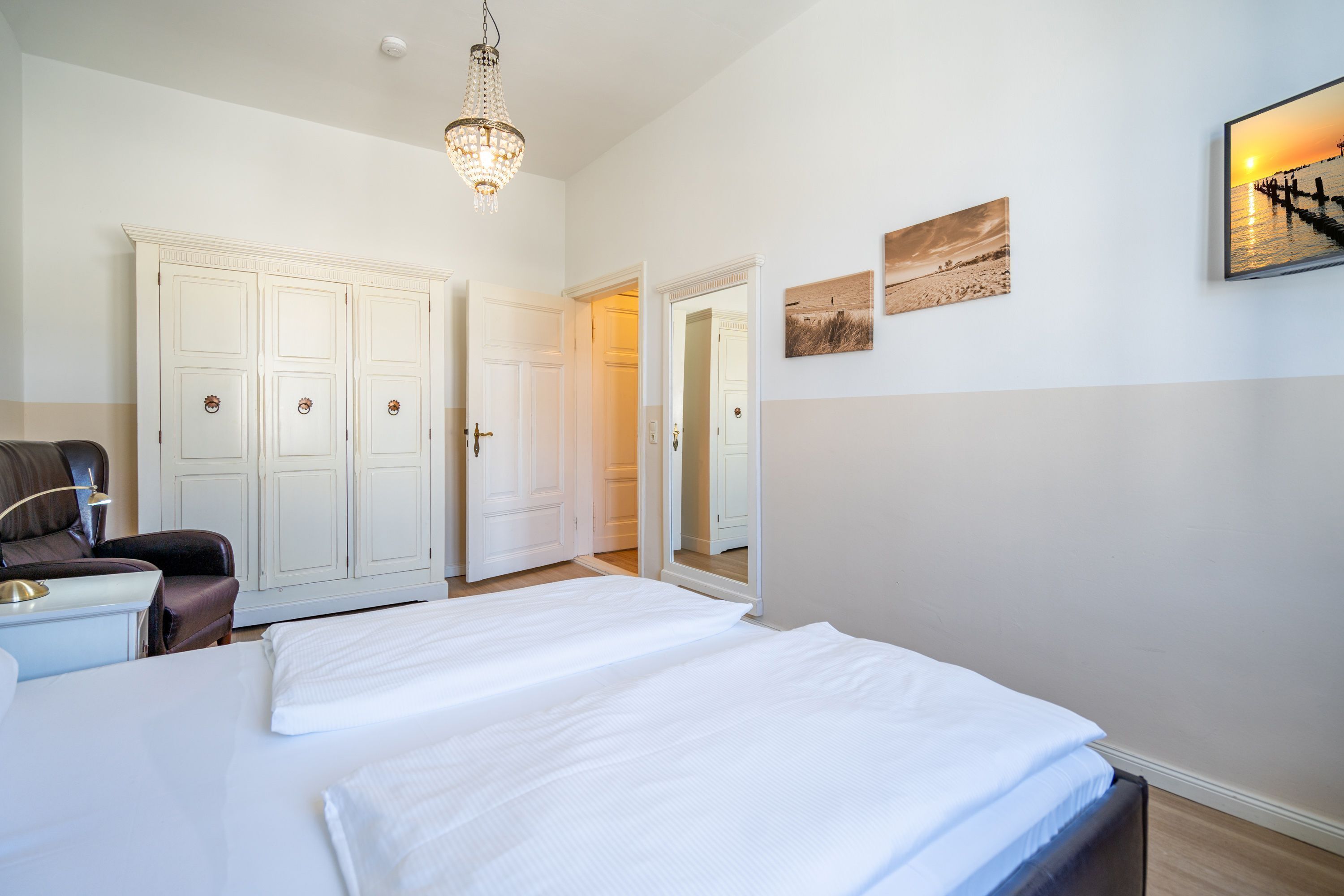 A bedroom with a large bed, a nightstand, a chair, and a wardrobe. There is a chandelier hanging from the ceiling and a painting on the wall.