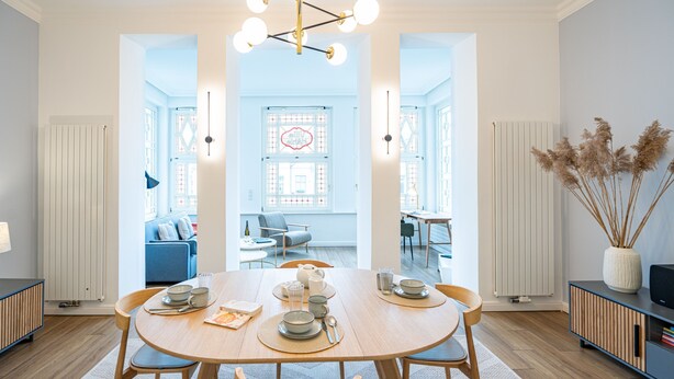 A dining area with a round table set for four, flanked by two chairs. A chandelier hangs above, and a vase with dried plants is on the right.