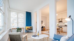 A modern living room with a sofa, armchair, and coffee table, a dining area with a table and chairs, and a kitchen with cabinets and appliances.
