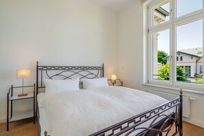 A neatly arranged bedroom with a bed, bedside lamps, a nightstand, and a window with a view of buildings. - Villa Hähle, App. 6, Germany (Heringsdorf)