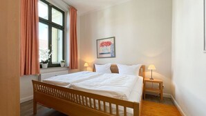 A bedroom with a wooden bed, two bedside lamps, a window with curtains, and a framed picture on the wall. - Villa Goodewind, App. 4 (Heringsdorf)