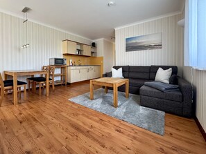 A compact living space with a wooden dining table, chairs, a gray sofa, and a kitchen area with a microwave and cabinets.