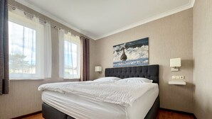 A bedroom with a large bed, two bedside lamps, a painting on the wall, and a window with curtains. - Maxine, Trassenheide (Baltic seaside resort), Germany (Trassenheide)
