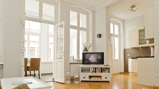 A spacious living room with a wooden floor, a television on a white entertainment unit, and a dining area with wicker chairs. - Schloss am Meer, App. 4 (Heringsdorf)