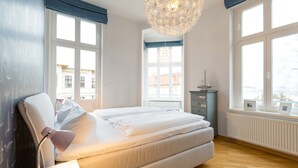 A modern bedroom with a large bed, a bedside table with a lamp, a dresser, and a view of the outside through large windows. - Schloss am Meer, App. 4 (Heringsdorf)