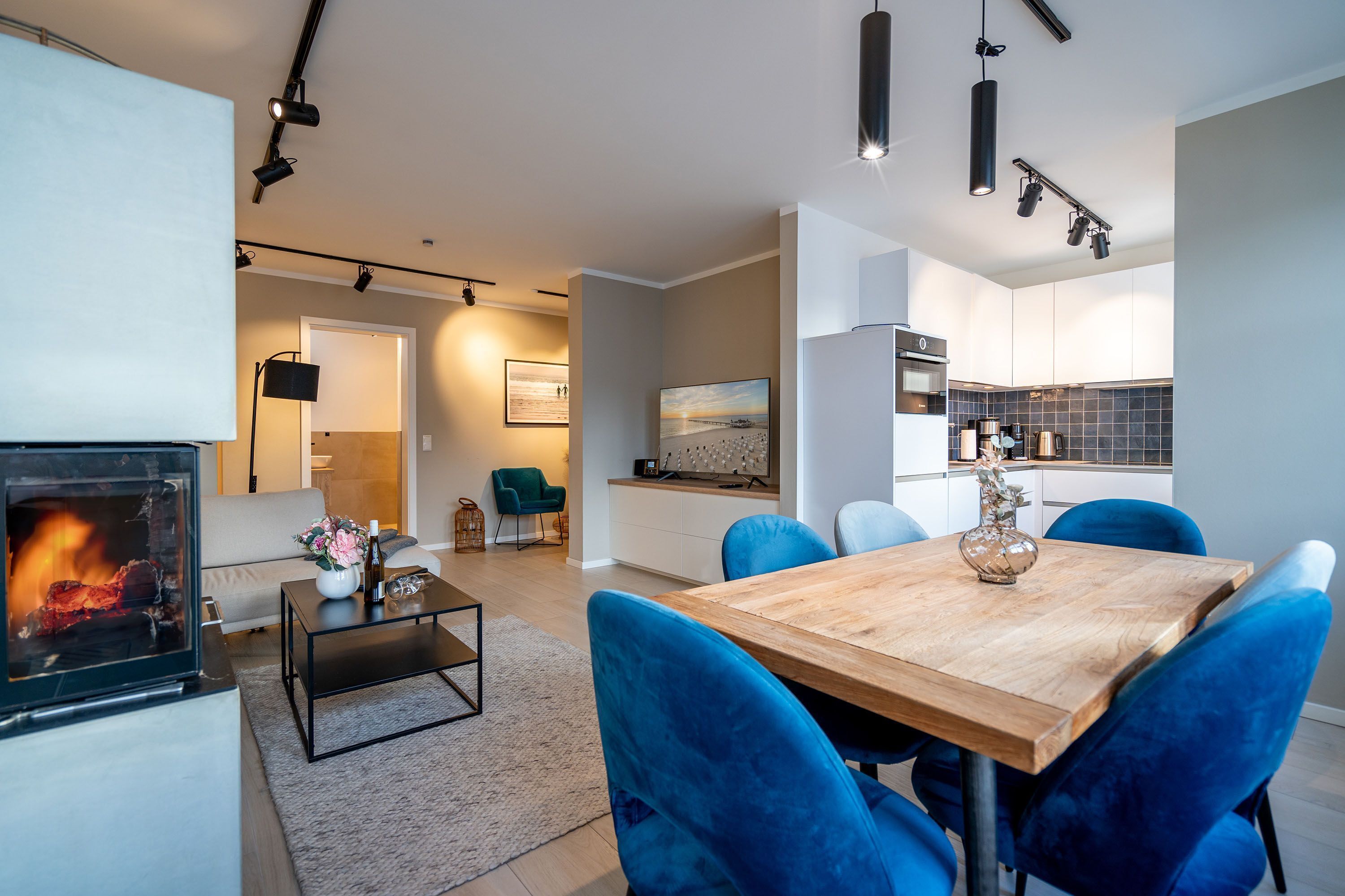 A modern living room with a fireplace, a dining area with blue chairs, and a kitchen with white cabinets.