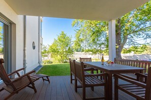 Une terrasse couverte avec une table et des chaises en bois, un banc et une vue sur une pelouse et des arbres.