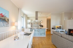 Une cuisine moderne avec un îlot central, un coin repas avec une table et des chaises blanches, un salon avec un canapé et une cheminée, et un tableau accroché au mur.