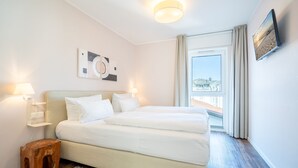 A hotel room with a bed, bedside lamps, a wooden stool, a rug, a painting, and a window with curtains. - Quartier Ahlbeck, app. 04 (Heringsdorf)